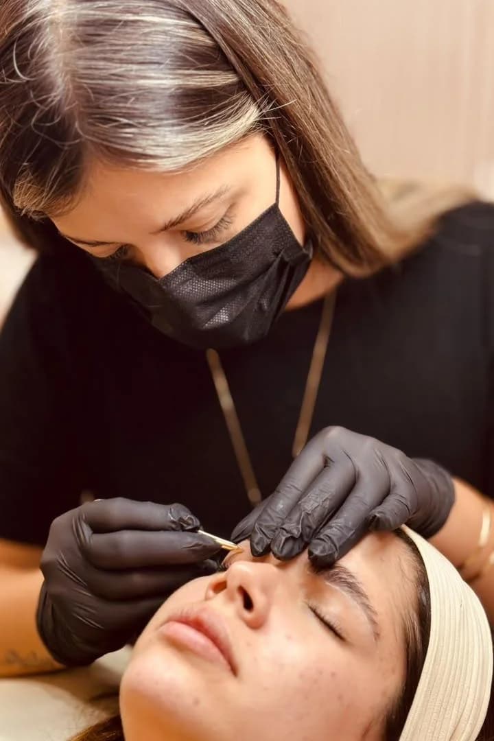 Amanda Farias concentrada, fazendo o design de sobrancelhas de uma cliente de olhos fechados.