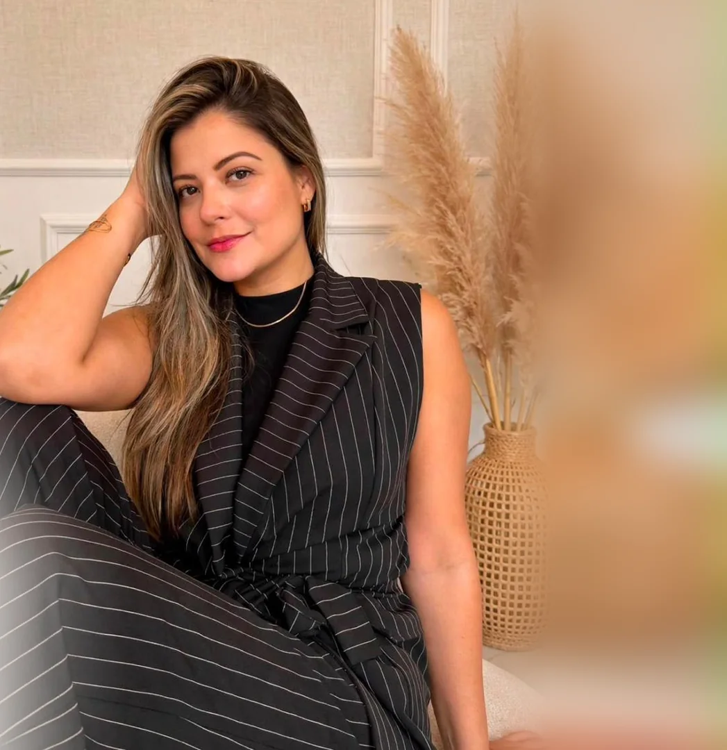 Amanda Farias sorrindo, sentada no seu estúdio de beleza, com a mão no cabelo, entre pampas e folhagens.